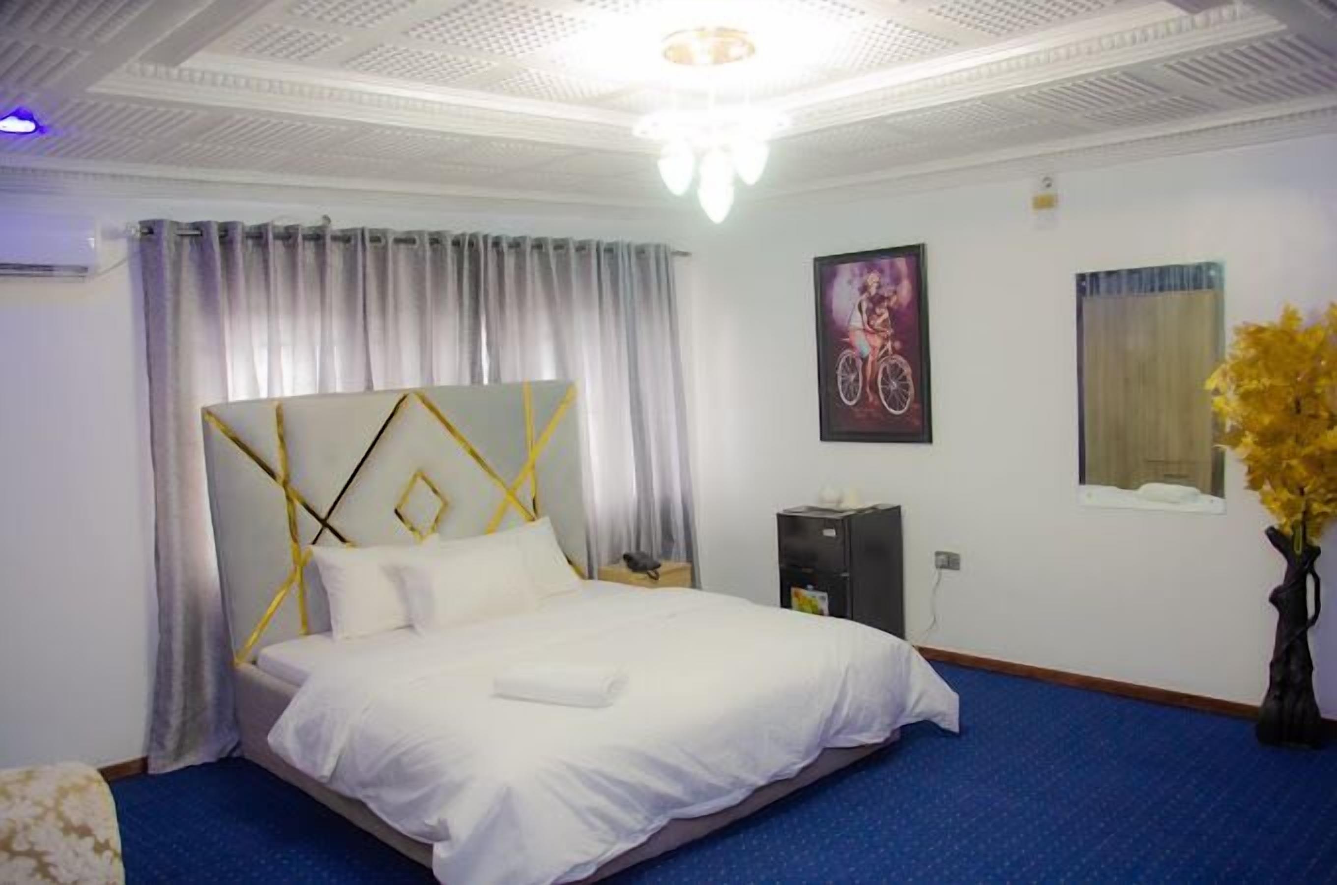 Una camera d'albergo con un letto, un comodino, una scrivania, una sedia, una finestra con tende, un vaso con fiori e un quadro appeso al muro.