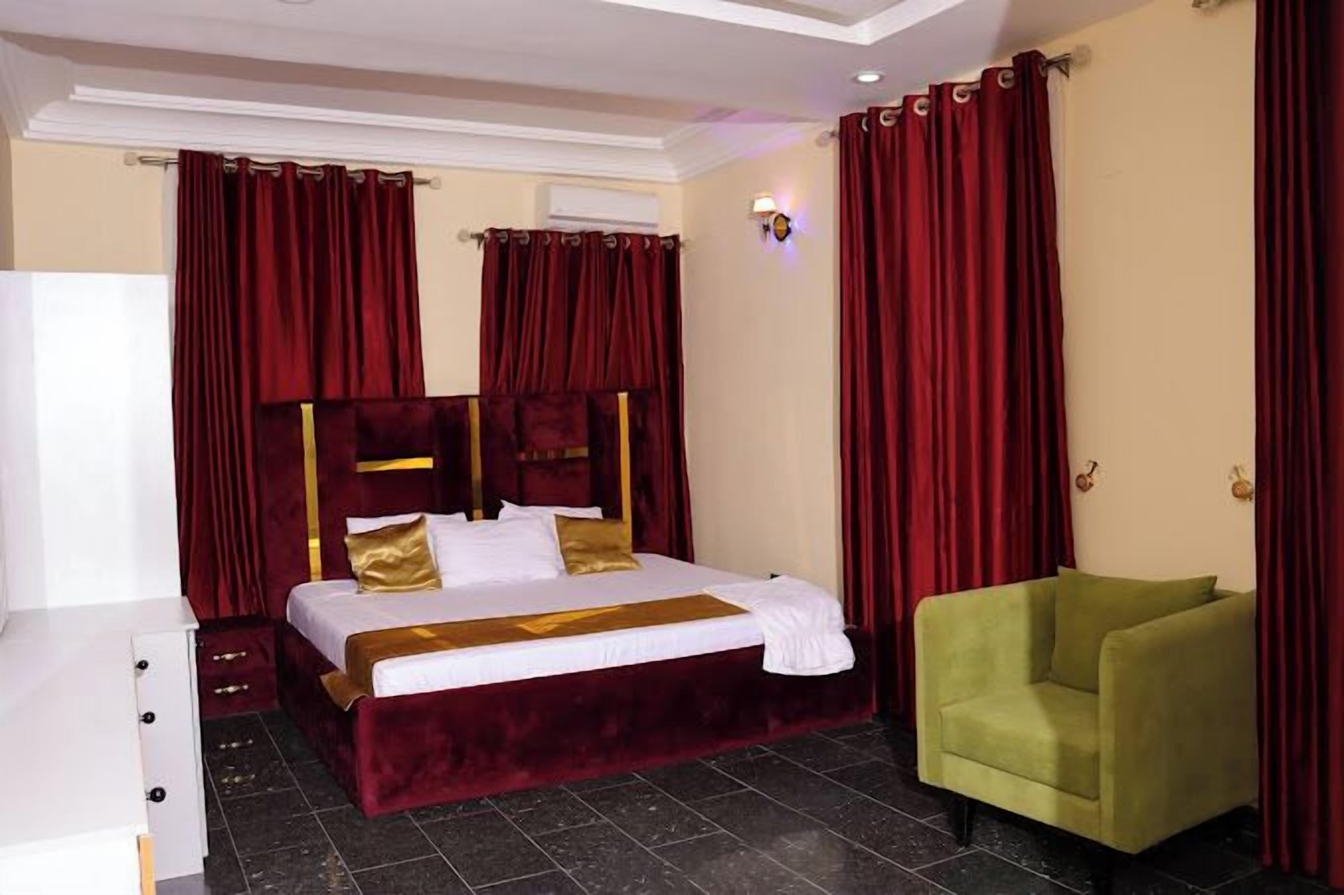 A hotel room with a large bed, red curtains, a green armchair, and a white dresser.