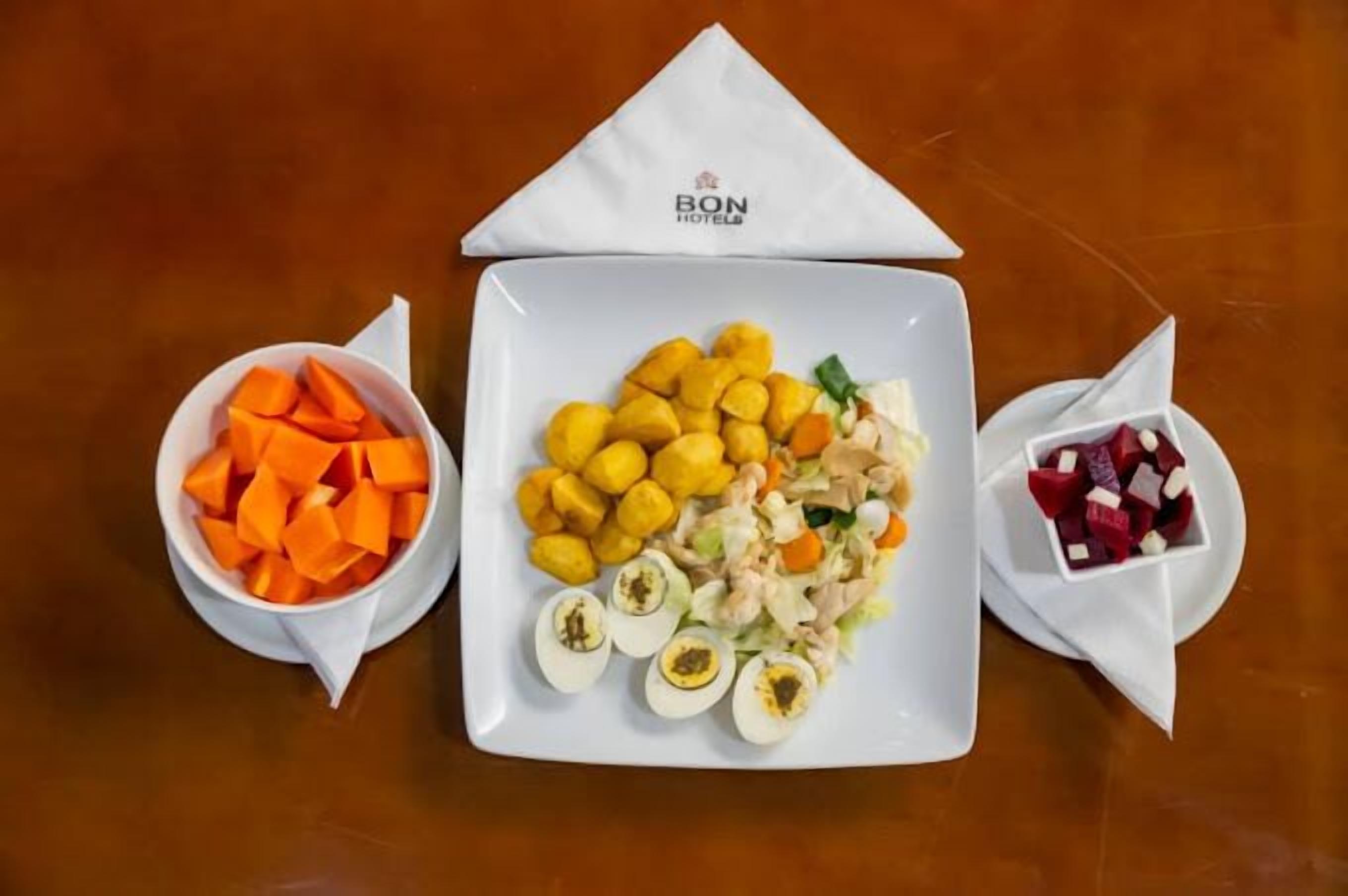 A square white plate with a salad, surrounded by three bowls of vegetables on a wooden table.