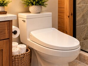 A toilet with a white lid, a roll of toilet paper, a basket with towels, and a potted plant on the counter. - NEW! Seneca 113: Private Entrance, Hot Tub, Heated Garage, Sleeps 6! (Snowshoe)