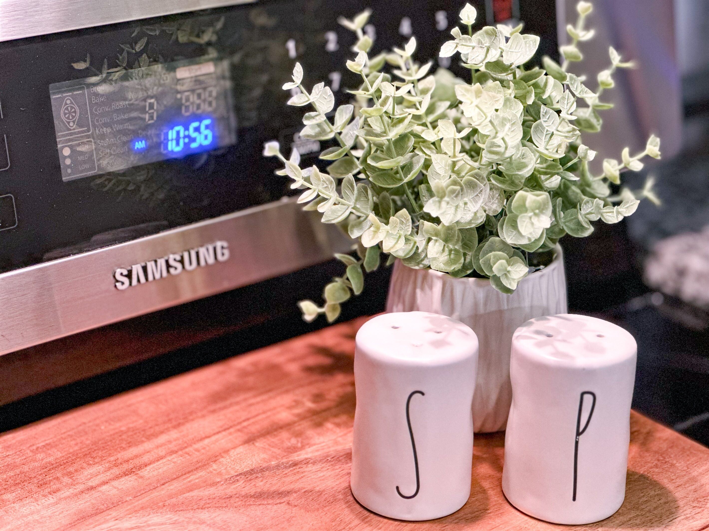 A Samsung microwave with a digital clock displaying 10:56, a small potted plant, and two salt and pepper shakers on a wooden surface.