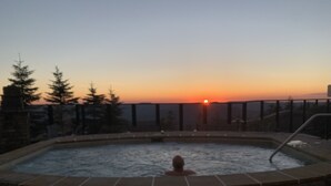 A hot tub with a stone border, surrounded by a stone wall and a metal railing, with a sunset in the background. - NEW! Seneca 113: Private Entrance, Hot Tub, Heated Garage, Sleeps 6! (Snowshoe)