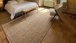 Un dormitorio con pared de piedra, una cama con ropa de cama blanca, un banco tejido, una silla, una lámpara de pie y una ventana con cortinas.