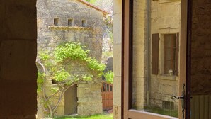 Una puerta abierta que da a un edificio de piedra con vistas a un jardín, una mesa con copas de vino y un jarrón verde, y un interior rústico.