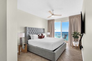 A spacious bedroom with a large bed, a ceiling fan, a balcony with a chair, and a TV mounted on the wall. - Deluxe Views-Short Walk 2 Beach-Resort Amenities (Miramar Beach)