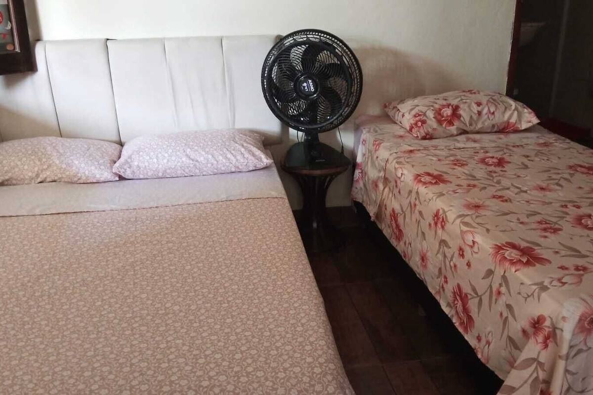 Une chambre avec deux lits, l’un recouvert d’une couvre-lit à motifs floraux et l’autre d’une simple couverture. Un ventilateur sur pied est placé entre les deux lits.