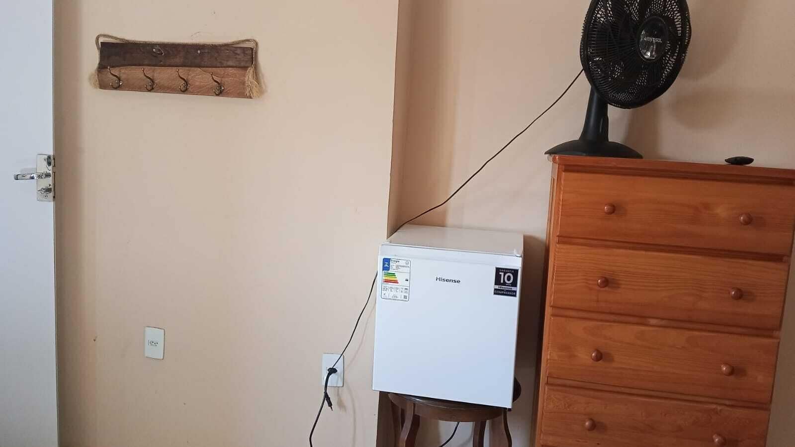A compact room with a white Hisense refrigerator, a wooden dresser, and a black fan on top.