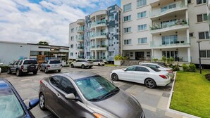 A multi-story residential building with balconies, a parking area with several cars, and a well-maintained lawn.
