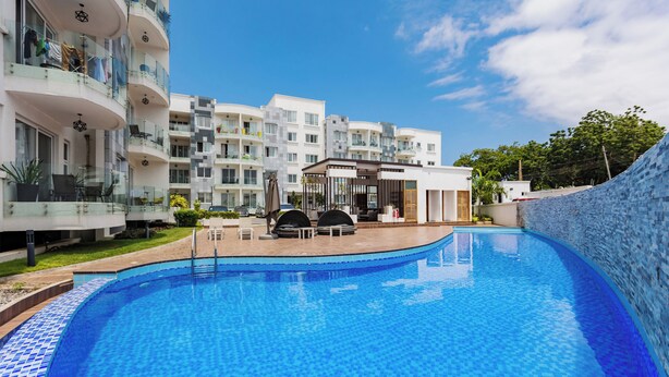 A swimming pool with a tiled edge, surrounded by a modern building with balconies and a covered seating area.