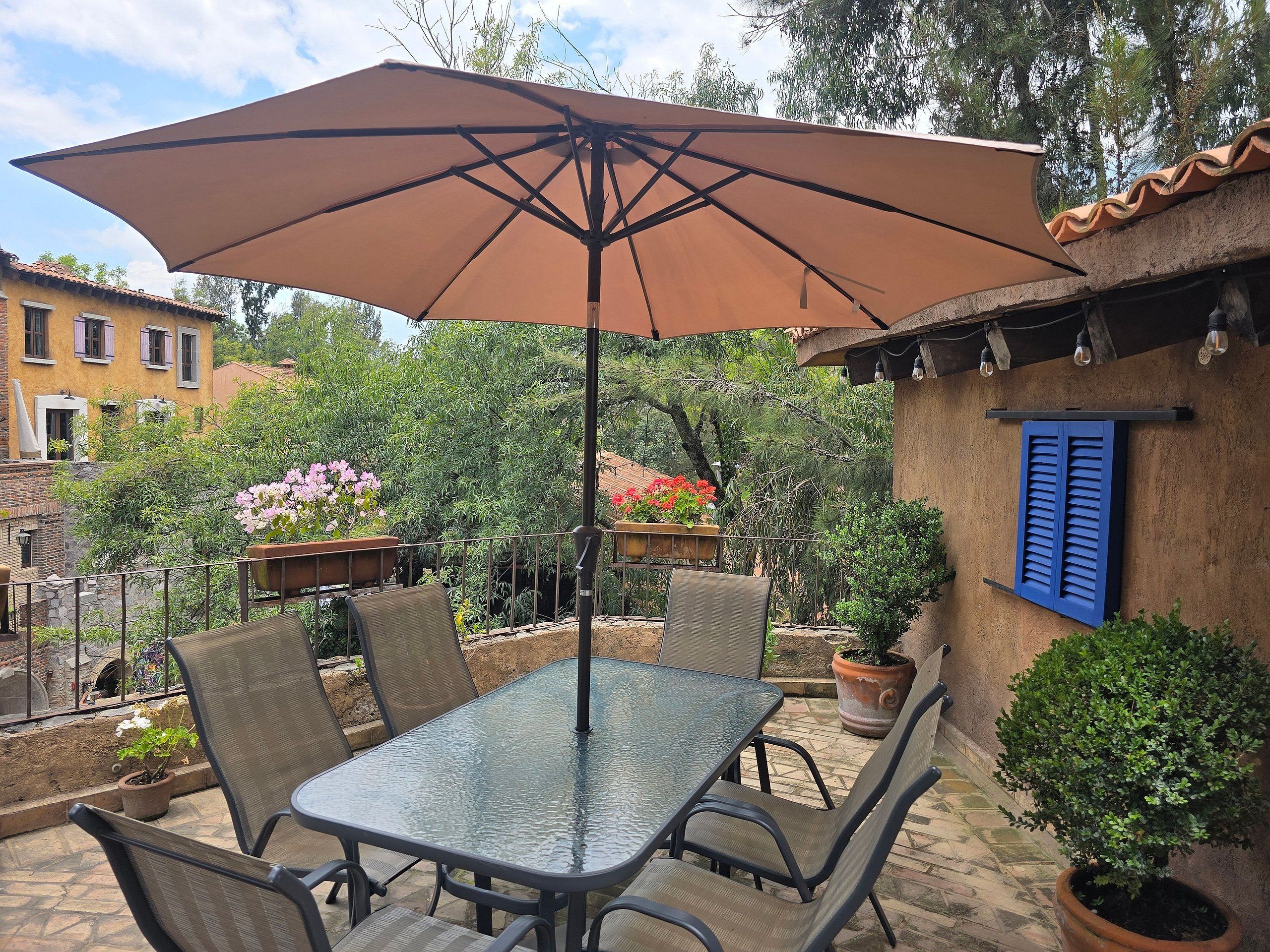 Un patio con una mesa y sillas de vidrio, una sombrilla y plantas en macetas.