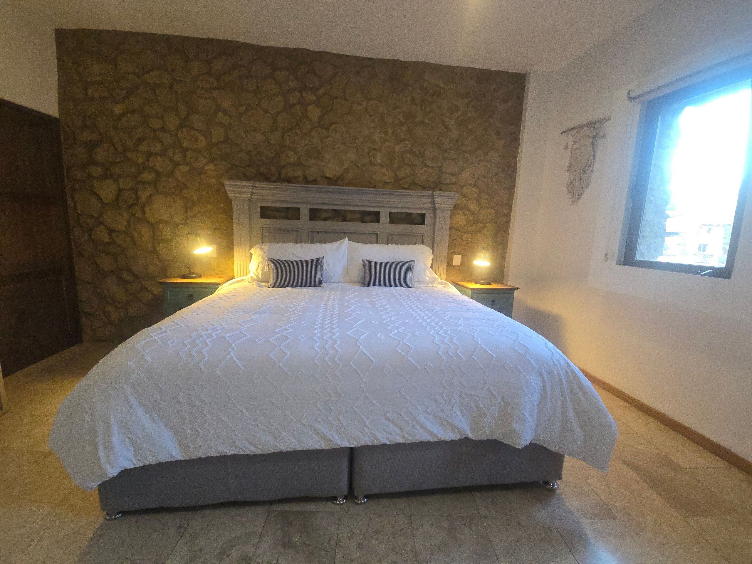 A bedroom with a stone wall, a bed with white bedding, two bedside tables with lamps, and a window.