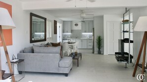 Een moderne woonkamer met een grijze bank, een salontafel en een tv-meubel. In de achtergrond bevindt zich een keuken met witte kasten en een plafondventilator.