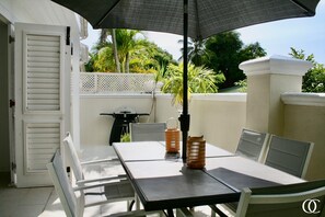 A patio with a table and chairs, an umbrella, and a white fence with greenery in the background.