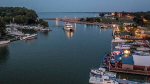 A marina with multiple boats docked, a lighthouse in the background, and a waterfront restaurant with outdoor seating. - “Blame Jaime” downtown, the heart of all the fun! (Port Clinton)