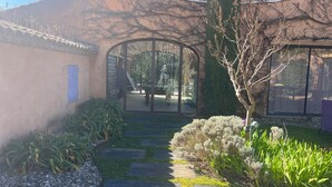 Un sendero conduce a un edificio con puerta de vidrio, rodeado de follaje y un jardín con baldosas de piedra.