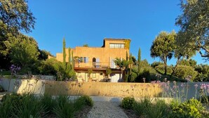 Una villa de estilo mediterráneo con un camino de grava que conduce a la entrada, rodeada de exuberante follaje y plantas con flores.