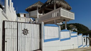 Ein Strandgrundstück mit weißer Fassade, einem Balkon mit Reetdach und einem eingezäunten Eingang mit Schiffsschwengel.
