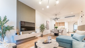 A modern living room with a flat-screen TV, a dining area, and a kitchen in the background.