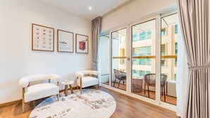 A modern living room with a large sliding glass door leading to a balcony, two white armchairs, a small round rug, and framed coffee-themed artwork on the wall. - Upgraded 2BR with Partial Sea and City Views Near the Beach, Elite Residence (Dubai)
