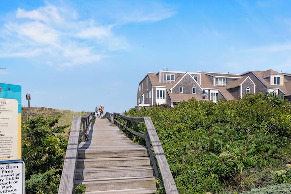 A wooden boardwalk leading to a row of houses with a sign on the left. - Ocean view spacious 5-bedroom Cape Cod style house with roof deck, AC, WiFi. (Plymouth)
