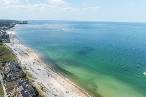 An aerial view of a beach with clear waters, a sandy shore, and buildings along the coastline. - Ocean view spacious 5-bedroom Cape Cod style house with roof deck, AC, WiFi. (Plymouth)