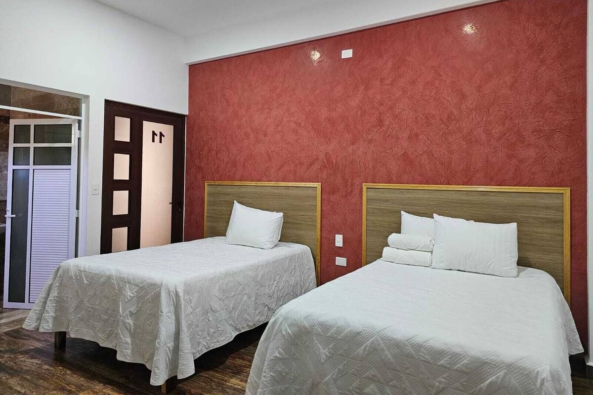 Habitación de hotel con dos camas, una pared roja con textura y una puerta de madera con panel de vidrio.