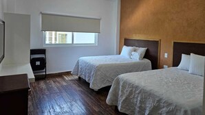 Ein Hotelzimmer mit zwei Betten, einem Fenster mit Jalousie, Holzboden und einem kleinen Schreibtisch mit Stuhl.