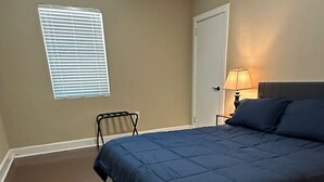 A bedroom with a bed, a ceiling fan, a window with blinds, a door, and a nightstand with a lamp. - The Greenfield: 2-bedroom townhome close to LSU (Baton Rouge)