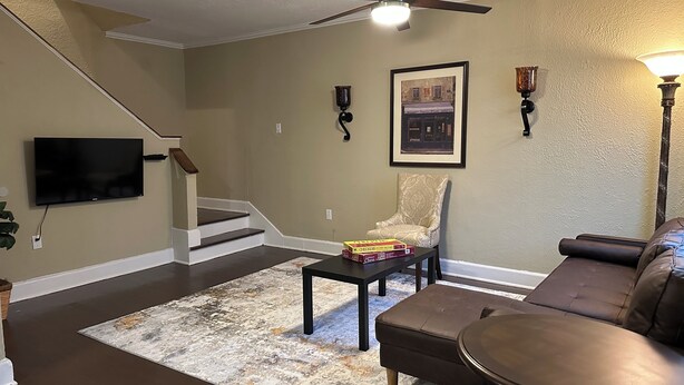 A living room with a staircase, a television, a patterned armchair, a brown leather sofa, and a chess set on a coffee table. - The Greenfield: 2-bedroom townhome close to LSU (Baton Rouge)