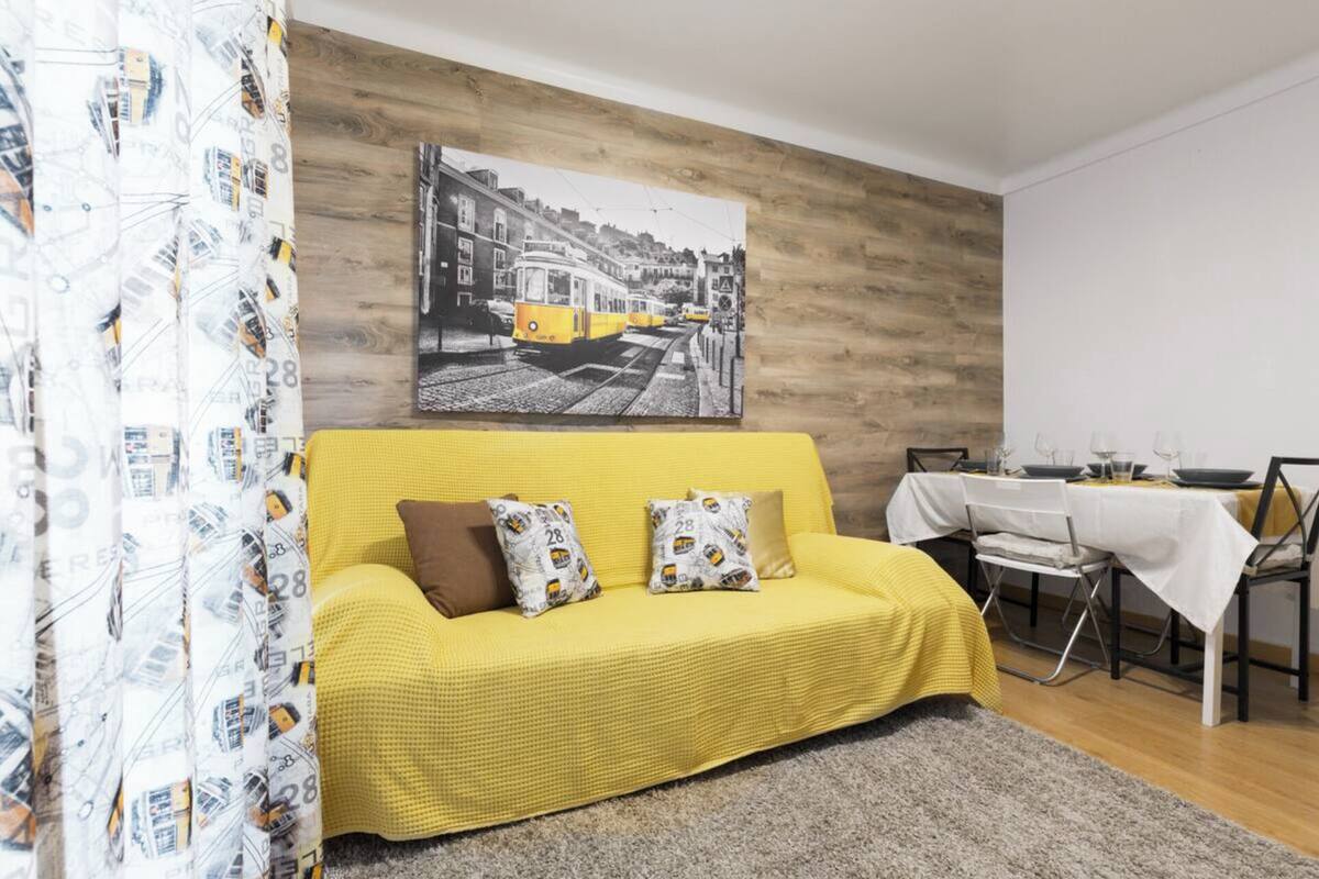 Un canapé jaune agrémenté de coussins à motifs, une table à manger avec des chaises et un tableau représentant un paysage urbain accroché au mur.