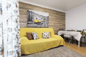 Een gele bank met gekleurde kussens, een eettafel met stoelen, en een stadsgezicht schilderij aan de muur.