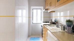 A kitchen with white tiles, wooden cabinets, a window, and a blue rug. - Alfama Vibes Apartments (Lisbon)
