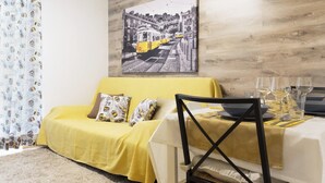 A yellow sofa with patterned cushions, a dining table set with a bowl and wine glasses, and a framed black and white photograph on the wall. - Alfama Vibes Apartments (Lisbon)