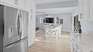 A modern kitchen with white cabinetry, a stainless steel refrigerator, a dining area with a white table and chairs, and a large window with a view of the outside.