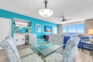 A modern living room with a glass dining table, patterned sofas, a kitchen with white cabinets, and a large window with a view.