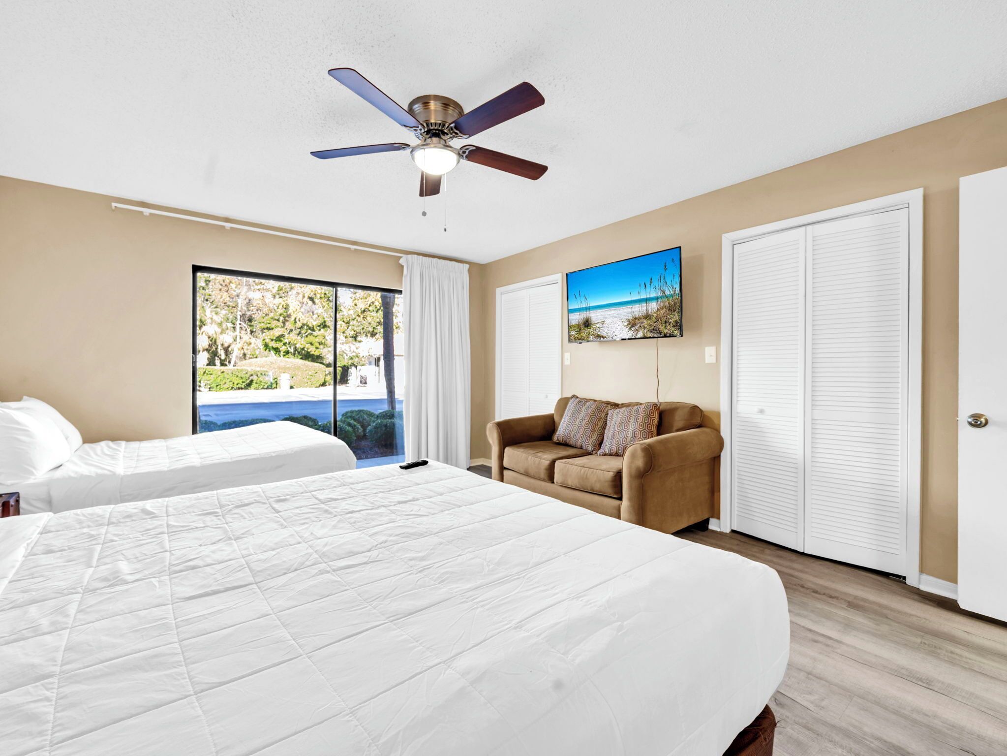 Habitación de hotel con dos camas, un ventilador de techo, un ventanal con vistas al exterior, un sofá marrón y un armario blanco.