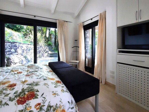 A bedroom with a bed, bench, TV, and a view of a garden and stone wall.