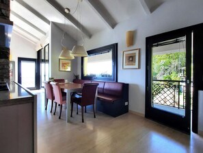 Un espace de repas moderne avec une table et des chaises en bois, un banc, des luminaires suspendus et de grandes fenêtres offrant une vue sur la verdure.