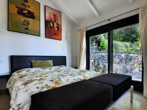 Une chambre à coucher avec un lit, une tête de lit, deux tableaux et une vue sur la verdure par une fenêtre.
