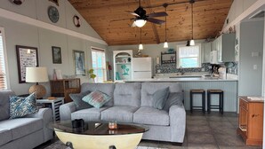 A living room with a grey sofa, a glass coffee table, a wooden ceiling, and a kitchen area with white cabinets and a tiled backsplash.