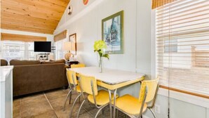 A cozy living area with a wooden ceiling, a dining table with yellow chairs, a brown sofa, and a TV on the left.