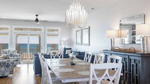 Een ruime woonkamer met een grote eettafel, witte stoelen en uitzicht op de oceaan door grote ramen.