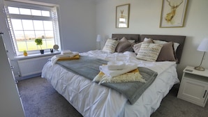 A neatly made bed with a grey blanket, a bedside table with a lamp, and a window with a view of the outdoors.