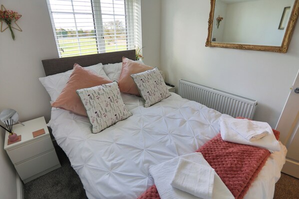 A neatly made bed with a white duvet and pink pillows, a bedside table with a lamp and a vase of flowers, and a window with blinds.