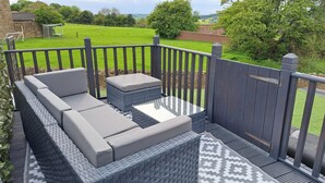 A deck with a sofa and ottoman, a patterned rug, and a view of a green field.