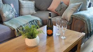 A wooden table with a bottle of Prosecco wine and two glasses, a potted plant, and a cozy living room with a sofa and pillows.