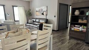 A dining area with a table and chairs, a sofa, a painting on the wall, and a bookshelf with items. - Mile-high Pines Lodge (South Lawrence)