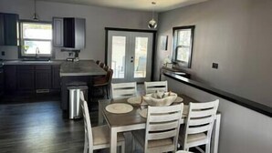 A modern kitchen with a dining area, featuring a central island, white chairs, and a large window. - Mile-high Pines Lodge (South Lawrence)