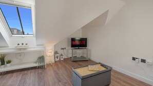 A modern loft bedroom with a skylight, a television displaying Netflix, a small bedside table, and a cozy seating area with cushions. - Trinity House Apartment | Grampian Lettings (Aberdeen)