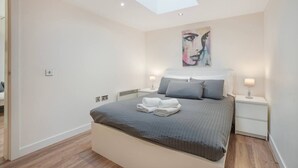 A modern bedroom with a skylight, a large bed with grey bedding, two bedside tables with lamps, and a painting on the wall. - Trinity House Apartment | Grampian Lettings (Aberdeen)